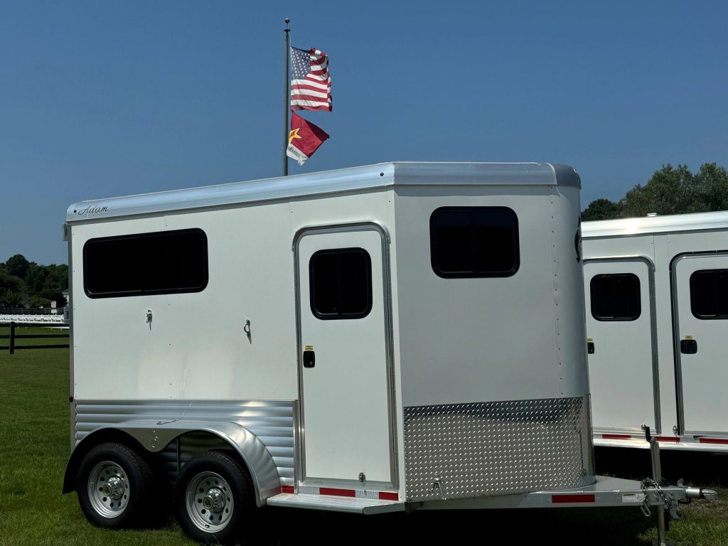 White Adam horse trailer parked on grass with American and state flags in the background.